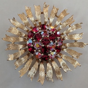 May include: A gold-toned metal brooch in a floral design. The center features a cluster of red and iridescent crystals, surrounded by a sunburst of textured gold ribbons. A vintage-inspired accessory.