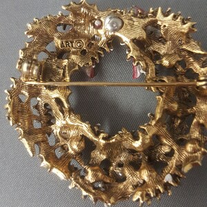 May include: Gold-toned metal Christmas wreath brooch with red and white enamel accents. The back of the brooch has a pin and a marking that reads "ARTG".