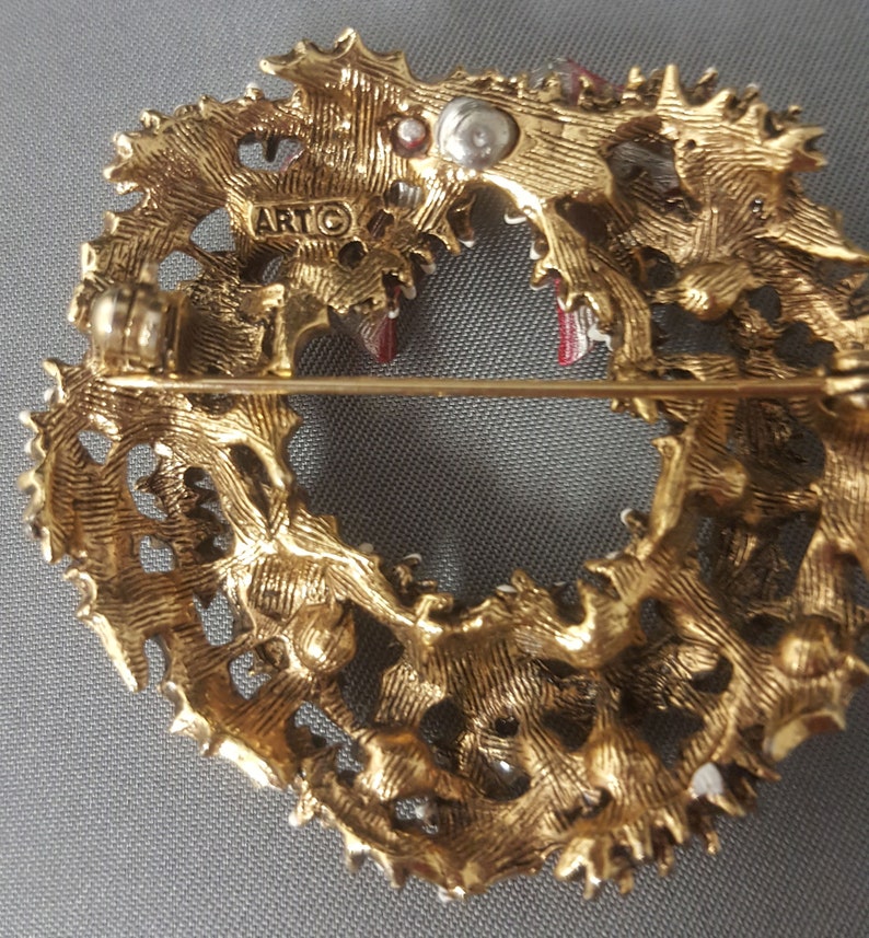 May include: Gold-toned metal Christmas wreath brooch with red and white enamel accents. The back of the brooch has a pin and a marking that reads "ARTG".