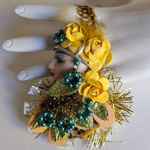 May include: A vintage brooch featuring a porcelain doll face with green and gold accents. The doll is wearing a yellow flower crown and a green and gold feathered headdress. The brooch is surrounded by yellow flowers and green beads.