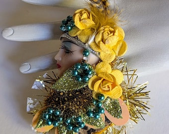 Puede incluir: Un broche vintage con un rostro de muñeca de porcelana con detalles en verde y dorado. La muñeca lleva una corona de flores amarillas y un tocado de plumas verde y dorado. El broche está rodeado de flores amarillas y cuentas verdes.
