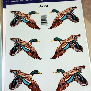 Puede incluir: Una hoja de cuatro calcoman&iacute;as pintadas a mano con patos coludos en vuelo. Los patos son marrones, verdes y azules con detalles blancos. Las calcoman&iacute;as est&aacute;n etiquetadas como "A-90" y "Calcoman&iacute;as pintadas a mano por Decorcal".