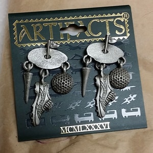 Peut inclure: Une paire de boucles d'oreilles en argent sur le thème du golf avec un charme de balle de golf, de chaussure de golf et de tee de golf. Les boucles d'oreilles sont sur une carte noire avec le mot "ARTIFACTS" et l'année "MCM LXXXVI" imprimés dessus.