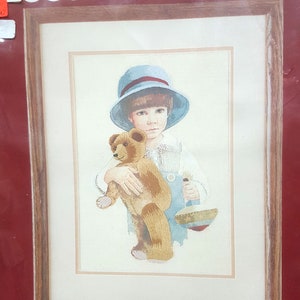 May include: A cross-stitch kit featuring a watercolor painting of a young child holding a teddy bear. The kit includes 100% polyester fabric, wool crewel yarn, cotton floss, needles, and instructions. The kit is made in the USA. The size of the finished piece is 10 inches by 10 inches.
