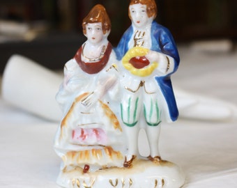 May include: A porcelain figurine of a couple in traditional clothing. The woman is wearing a white dress with red and gold accents, and the man is wearing a blue jacket with gold trim and green pants. The figurine is on a white base with gold accents.