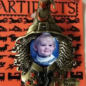 May include: A bronze-colored witch photo frame with a crescent moon and stars. The frame has a space for a 2x3 inch photo. The frame is on a red background with the word "ARTIFACTS" printed on it.