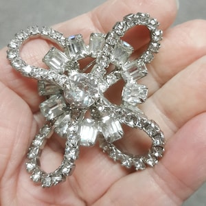 Könnte beinhalten: Eine silberfarbene Brosche mit einem großen, klaren Strassstein in der Mitte und kleineren Strasssteinen, die ihn umgeben. Die Brosche ist in Form einer Schleife mit zwei Schlaufen gestaltet.