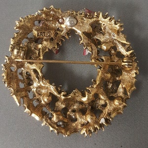 May include: Gold-tone metal Christmas wreath brooch with red and white enamel accents. The back of the brooch has a pin and a clasp, and the word "ARTC" is stamped on the back.