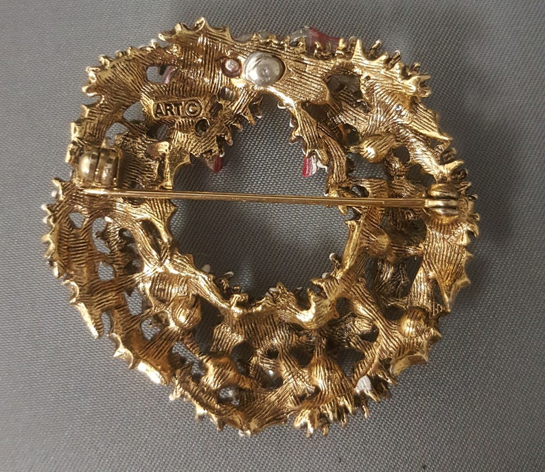 May include: Gold-tone metal Christmas wreath brooch with red and white enamel accents. The back of the brooch has a pin and a clasp, and the word "ARTC" is stamped on the back.