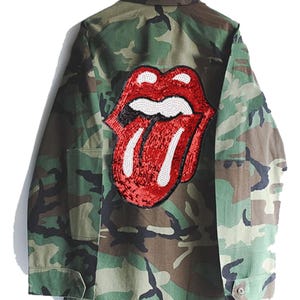Puede incluir: Chaqueta verde camuflaje con un parche de lengua y labios rojos y negros con lentejuelas en la espalda.