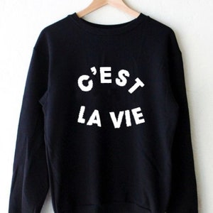 Puede incluir: Sudadera negra con texto blanco que dice "C'est la vie" en la parte delantera.