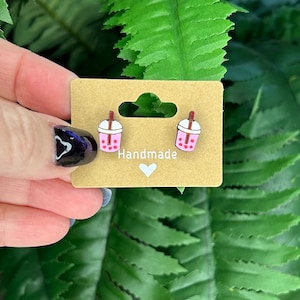 May include: A pair of handmade pink bubble tea earrings with a brown straw and white lid. The earrings are on a brown card with the word "Handmade" and a white heart.