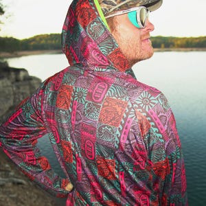 May include: A hooded long-sleeve shirt with a vibrant, colorful pattern of pink, orange, teal, and purple geometric shapes. The person is wearing a camouflage baseball cap and sunglasses. The background shows a lake and trees.
