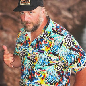 May include: Black cap with a patch featuring a mountain and tree design. A short-sleeved button-down shirt with a colorful wave and surfboard pattern. The shirt has a black collar and a small logo patch.