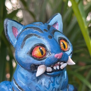 Peut inclure: Figurine de chat en c&eacute;ramique bleue avec des rayures noires et de grandes dents. Le chat a des yeux orange et jaunes, des oreilles roses et un nez rose. La figurine est d&eacute;taill&eacute;e avec des taches et des rayures noires.