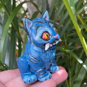 Peut inclure: Figurine bleue d'un chat stylis&eacute; avec des yeux orange et des dents pointues. Le chat a des rayures noires et est assis droit. La figurine est tenue dans la main d'une personne, avec un feuillage vert en arri&egrave;re-plan.