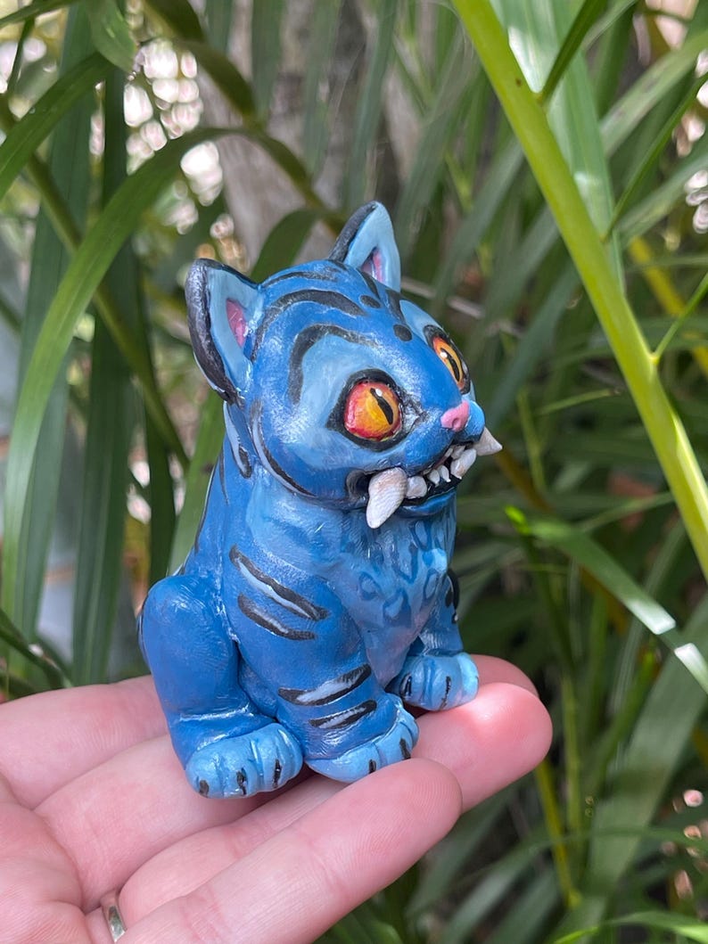 Peut inclure: Figurine bleue d'un chat stylis&eacute; avec des yeux orange et des dents pointues. Le chat a des rayures noires et est assis droit. La figurine est tenue dans la main d'une personne, avec un feuillage vert en arri&egrave;re-plan.