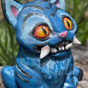 Peut inclure: Figurine de chat en c&eacute;ramique bleue avec de grands yeux orange, des dents pointues et des rayures noires. Le chat a un nez et des oreilles roses, et une surface textur&eacute;e d&eacute;taill&eacute;e. Une pi&egrave;ce d'art unique et artisanale.