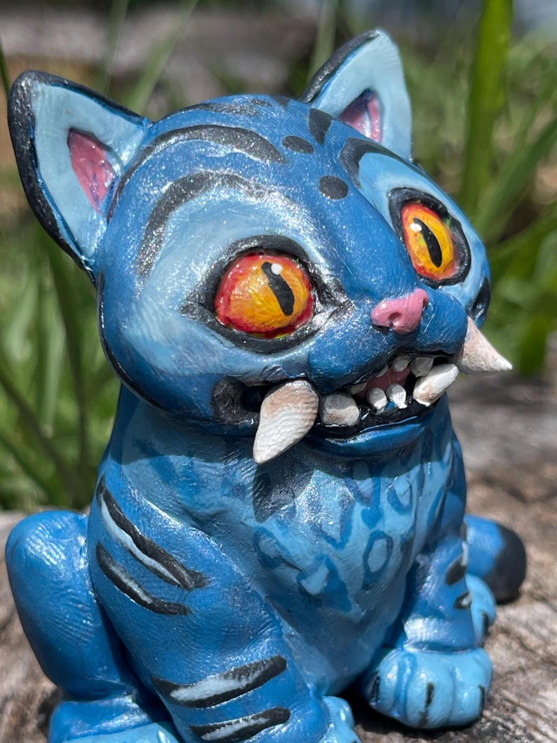 Peut inclure: Figurine de chat en c&eacute;ramique bleue avec de grands yeux orange, des dents pointues et des rayures noires. Le chat a un nez et des oreilles roses, et une surface textur&eacute;e d&eacute;taill&eacute;e. Une pi&egrave;ce d'art unique et artisanale.