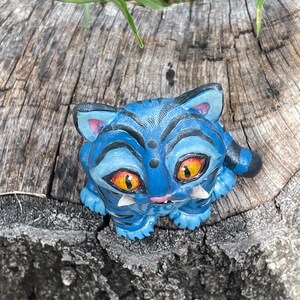 Peut inclure: Figurine bleue repr&eacute;sentant un chat avec des rayures noires et de grands yeux orange. Le chat a des crocs blancs et est pos&eacute; sur une surface en bois vieilli. Les oreilles du chat sont roses &agrave; l'int&eacute;rieur.