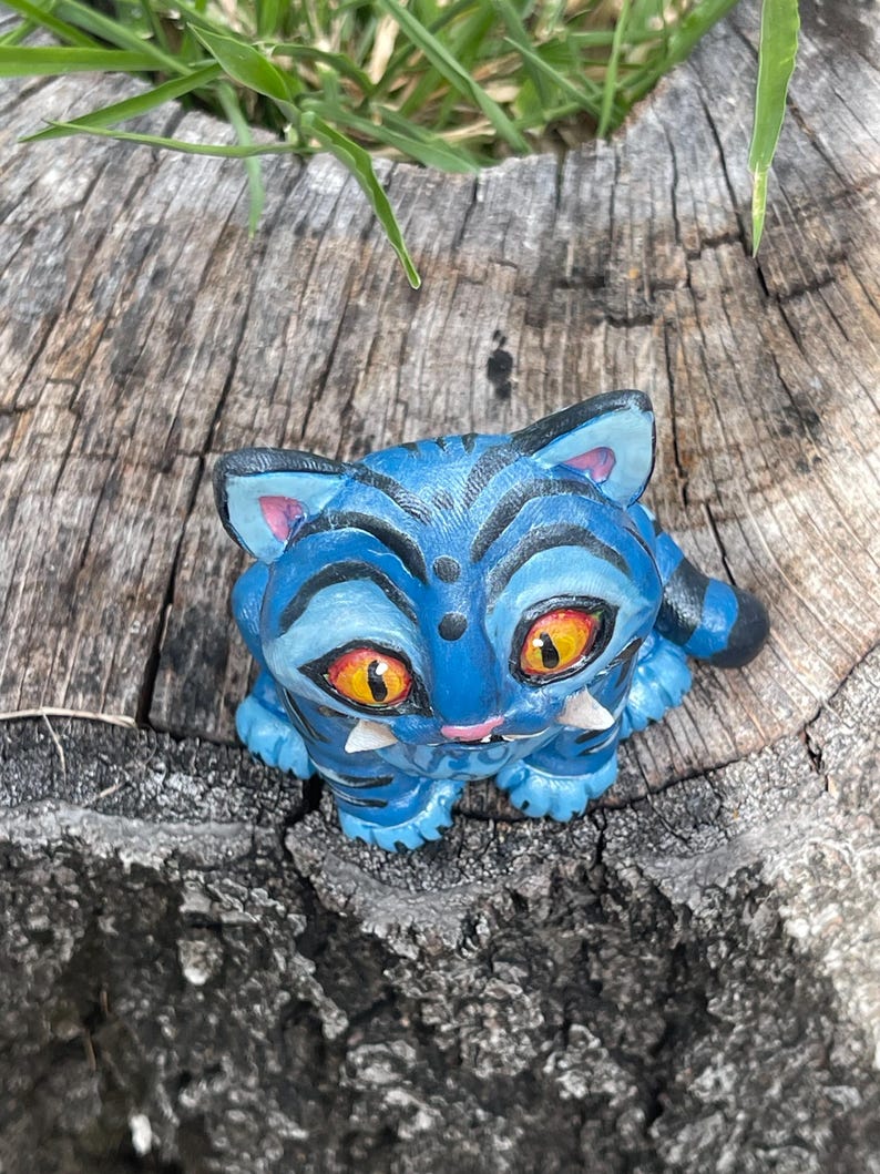 Peut inclure: Figurine bleue repr&eacute;sentant un chat avec des rayures noires et de grands yeux orange. Le chat a des crocs blancs et est pos&eacute; sur une surface en bois vieilli. Les oreilles du chat sont roses &agrave; l'int&eacute;rieur.