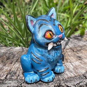 Peut inclure: Figurine de chat en c&eacute;ramique bleue avec des yeux orange et de grandes canines. Le chat a des rayures noires et est assis sur une surface en bois vieilli. La figurine a une apparence de dessin anim&eacute;.
