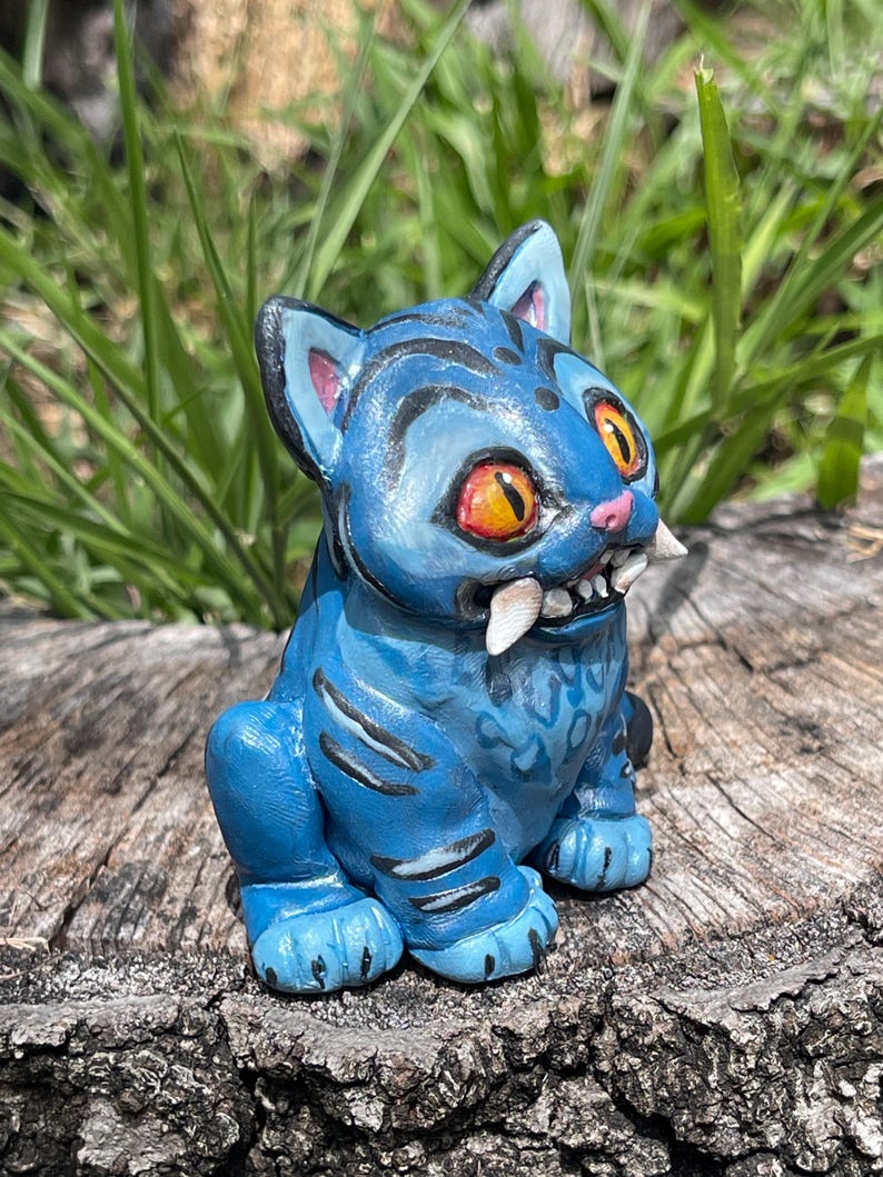 Peut inclure: Figurine de chat en c&eacute;ramique bleue avec des yeux orange et de grandes canines. Le chat a des rayures noires et est assis sur une surface en bois vieilli. La figurine a une apparence de dessin anim&eacute;.
