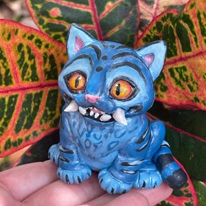 Peut inclure: Figurine de chat bleu avec des rayures noires, de grands yeux orange et des crocs pro&eacute;minents. Le chat est assis, les pattes visibles. Le fond pr&eacute;sente des feuilles color&eacute;es aux teintes rouges, vertes et jaunes. La figurine est tenue dans une main.