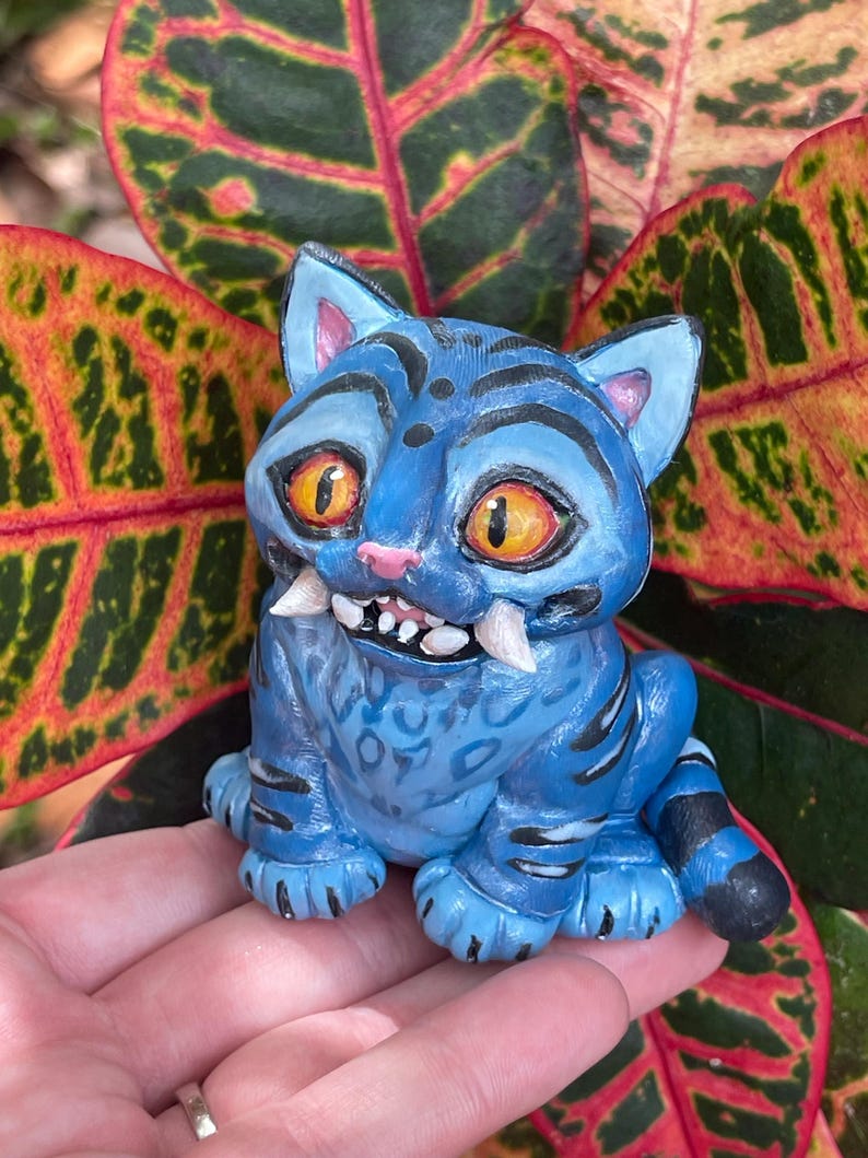 Peut inclure: Figurine de chat bleu avec des rayures noires, de grands yeux orange et des crocs pro&eacute;minents. Le chat est assis, les pattes visibles. Le fond pr&eacute;sente des feuilles color&eacute;es aux teintes rouges, vertes et jaunes. La figurine est tenue dans une main.