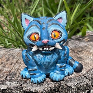 Peut inclure: Figurine de chat bleu avec des yeux orange, des crocs blancs et des rayures noires. Le chat est assis sur une souche d'arbre. La figurine a un design fantaisiste et de dessin anim&eacute;.
