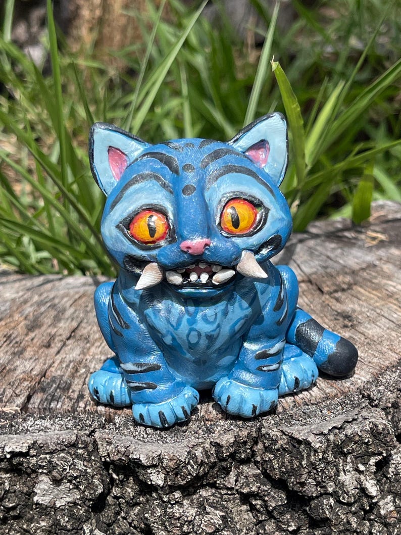 Peut inclure: Figurine de chat bleu avec des yeux orange, des crocs blancs et des rayures noires. Le chat est assis sur une souche d'arbre. La figurine a un design fantaisiste et de dessin anim&eacute;.