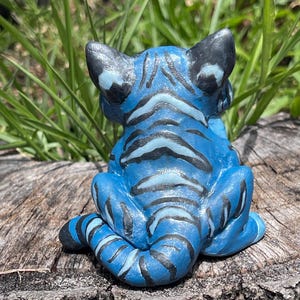 Peut inclure: Figurine de tigre bleu avec des rayures et des accents noirs. La sculpture en c&eacute;ramique est assise sur une souche d'arbre, le dos du tigre faisant face au spectateur. Les oreilles et la queue sont noires, et les rayures sont d'une nuance de bleu plus fonc&eacute;e.