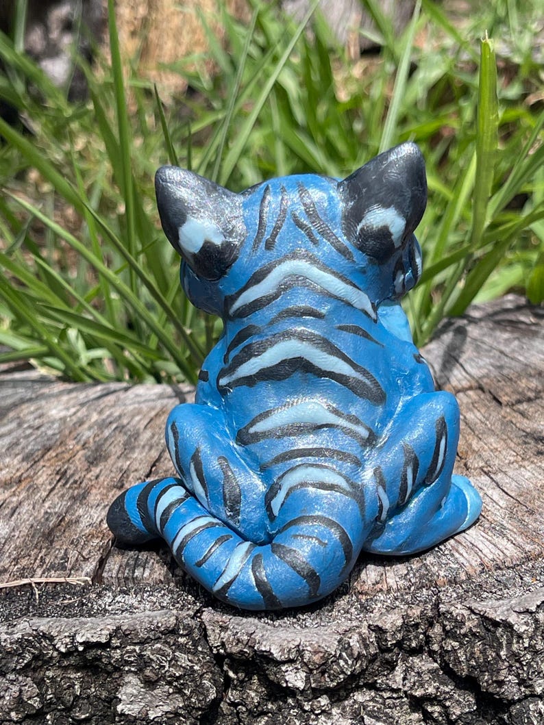 Peut inclure: Figurine de tigre bleu avec des rayures et des accents noirs. La sculpture en c&eacute;ramique est assise sur une souche d'arbre, le dos du tigre faisant face au spectateur. Les oreilles et la queue sont noires, et les rayures sont d'une nuance de bleu plus fonc&eacute;e.