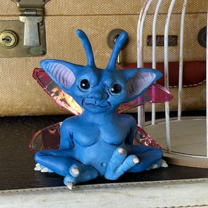 May include: A blue fairy figurine with iridescent wings, sitting with its legs crossed. The fairy has large eyes and a whimsical expression.