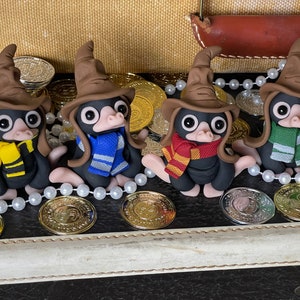 Peut inclure: Quatre figurines faites à la main de créatures Niffler de la série Harry Potter. Chaque Niffler porte un chapeau de sorcier brun et une écharpe de couleur différente. Les écharpes sont jaune et noire, bleu et blanche, rouge et or, et verte et blanche.