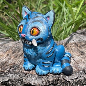 Peut inclure: Figurine de chat en c&eacute;ramique bleue avec des rayures noires, de grands yeux orange et des crocs. Le chat est assis sur une souche d'arbre, avec de l'herbe verte en arri&egrave;re-plan. La figurine a un design d&eacute;taill&eacute; et une expression enjou&eacute;e.