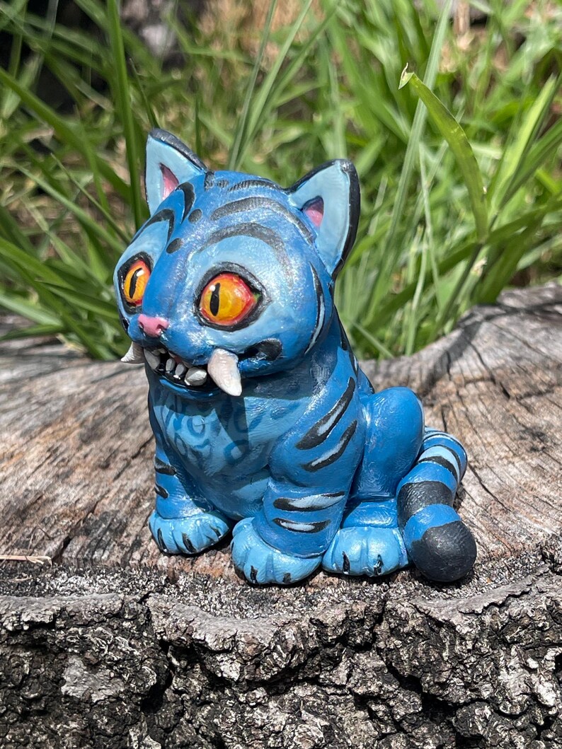 Peut inclure: Figurine de chat en c&eacute;ramique bleue avec des rayures noires, de grands yeux orange et des crocs. Le chat est assis sur une souche d'arbre, avec de l'herbe verte en arri&egrave;re-plan. La figurine a un design d&eacute;taill&eacute; et une expression enjou&eacute;e.