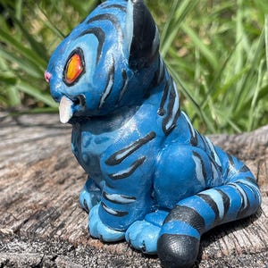 Peut inclure: Figurine bleue d'une cr&eacute;ature f&eacute;line avec des rayures noires. La figurine a des yeux orange, un nez rose et une dent blanche. La cr&eacute;ature est assise sur un morceau de bois, avec de l'herbe verte en arri&egrave;re-plan.