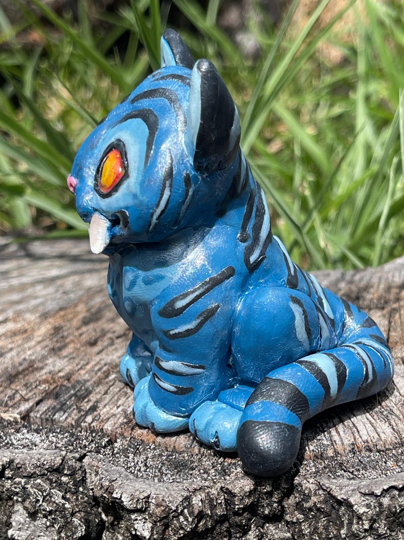 Peut inclure: Figurine bleue d'une cr&eacute;ature f&eacute;line avec des rayures noires. La figurine a des yeux orange, un nez rose et une dent blanche. La cr&eacute;ature est assise sur un morceau de bois, avec de l'herbe verte en arri&egrave;re-plan.