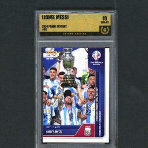 May include: A graded Panini Instant card featuring Lionel Messi. The card shows Messi holding a trophy with his team, celebrating a victory. The card is graded 10 GEM-MT by Golden Grading, with the text "COPA AMERICA USA 2021" and "ARGENTINA 1-0 COLOMBIA".