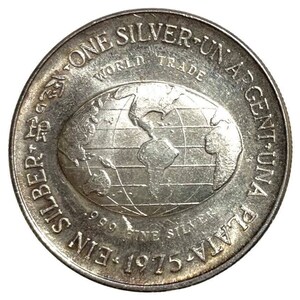 May include: A silver coin with a globe design and the text "EIN SILBER - 999 FINE SILVER - PLATA-UNA ARGENT-UN WORLD TRADE 1975" around the edge.