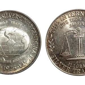 May include: Silver coin with a globe on one side and a scale on the other. The coin reads "One Silver-Un Argentuna Plata-S61-Ein 1975-1975" on one side and "The International Silver Trade One Troy Ounce .999 Fine 81.1 Grams" on the other.