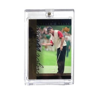 Puede incluir: Una tarjeta de intercambio de Tiger's Tales con una foto a color de Tiger Woods poniendo en un campo de golf. La tarjeta tiene un fondo dorado con el texto "Tiger Woods" en escritura negra. La tarjeta está en una caja protectora de plástico transparente.