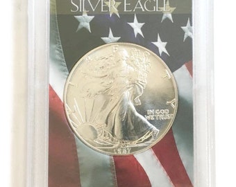 Könnte beinhalten: Eine silberne American Silver Eagle Münze in einer Schutzhülle. Die Münze zeigt ein Motiv der wandernden Lady Liberty und den Text "IN GOD WE TRUST" sowie das Jahr 1987. Der Hintergrund zeigt die amerikanische Flagge mit dem Text "AMERICAN SILVER EAGLE".