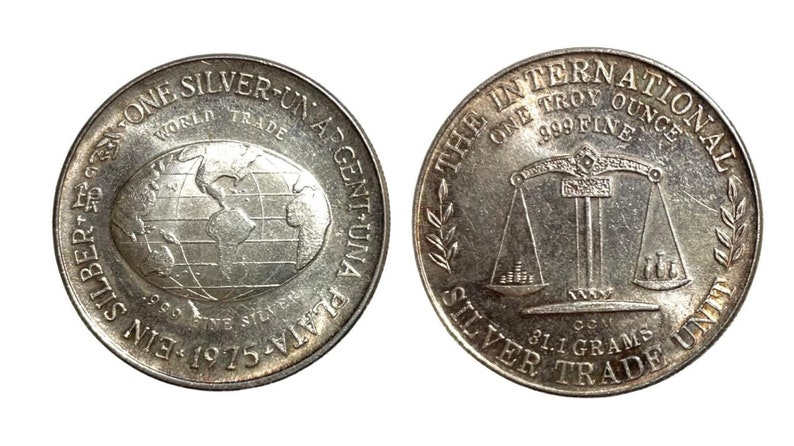 May include: Silver coin with a globe on one side and a scale on the other. The coin reads "One Silver-Un Argentuna Plata-S61-Ein 1975-1975" on one side and "The International Silver Trade One Troy Ounce .999 Fine 81.1 Grams" on the other.