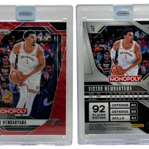 Op de afbeelding: Twee Victor Wembanyama basketbal trading cards in beschermende hoesjes. De voorkant heeft een rode achtergrond met de tekst "MONOPOLY" en "SAN ANTONIO SPURS". De achterkant toont spelersstatistieken en een spelerbeoordeling van "92".