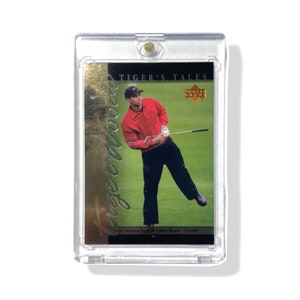 Puede incluir: Una tarjeta de golf de Tiger Woods de la serie Tiger's Tales. La tarjeta presenta una foto de Tiger Woods con una camisa roja y pantalones negros, balanceando un palo de golf. La tarjeta está en una funda protectora de plástico transparente.