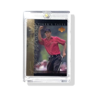 Puede incluir: Una tarjeta coleccionable de Tiger Woods en una funda protectora transparente. La tarjeta muestra a Woods con una camisa roja y gorra negra, celebrando una victoria. El texto en la tarjeta dice "TIGER'S TALES" y "Three straight Amateur wins for Tiger 1996."