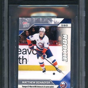 May include: A 2025-26 Topps Now Matthew Schaefer hockey sticker, graded 9.5 Gem Mint by Beckett. The sticker features the player in a white, blue, and orange New York Islanders uniform, holding a hockey stick on the ice, with the text Rookie included.