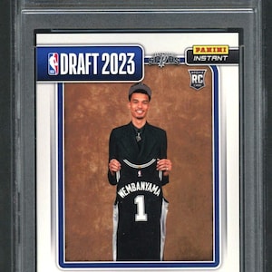 May include: A graded 2023 Panini Instant Draft Night Victor Wembanyama rookie card, numbered #DN1. The card features a photograph of the athlete holding a black San Antonio Spurs jersey with the number 1, encased in a protective plastic slab.